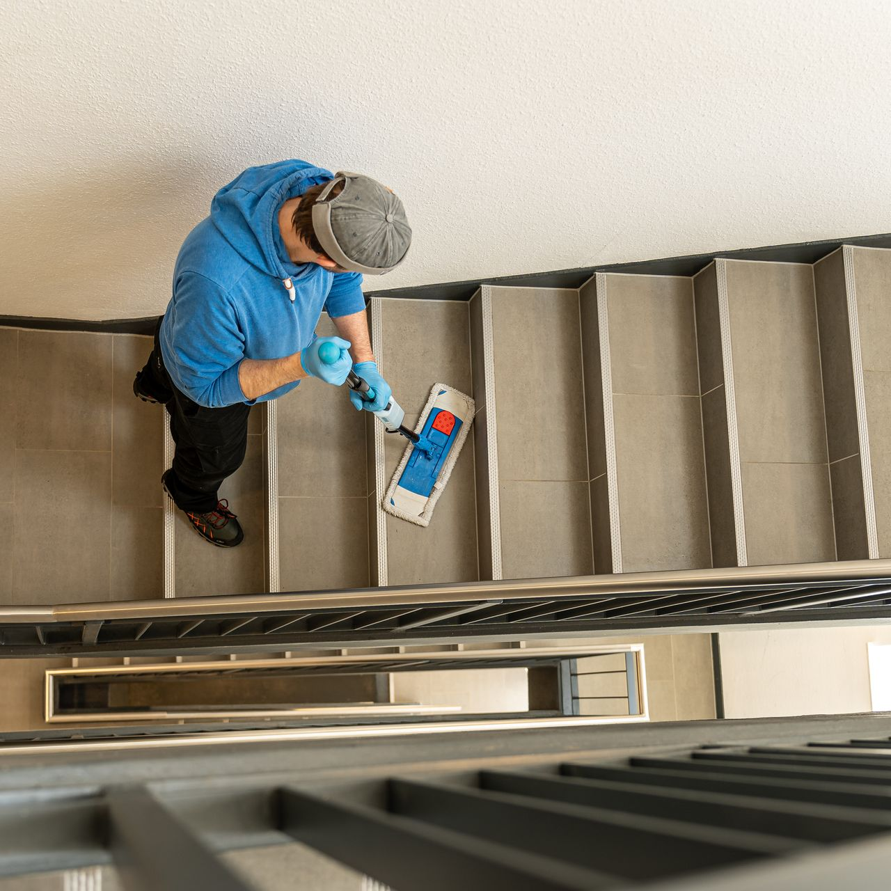 Vue d'en haut, une personne portant un sweat à capuche bleu et un bonnet gris, en train de laver un escalier.