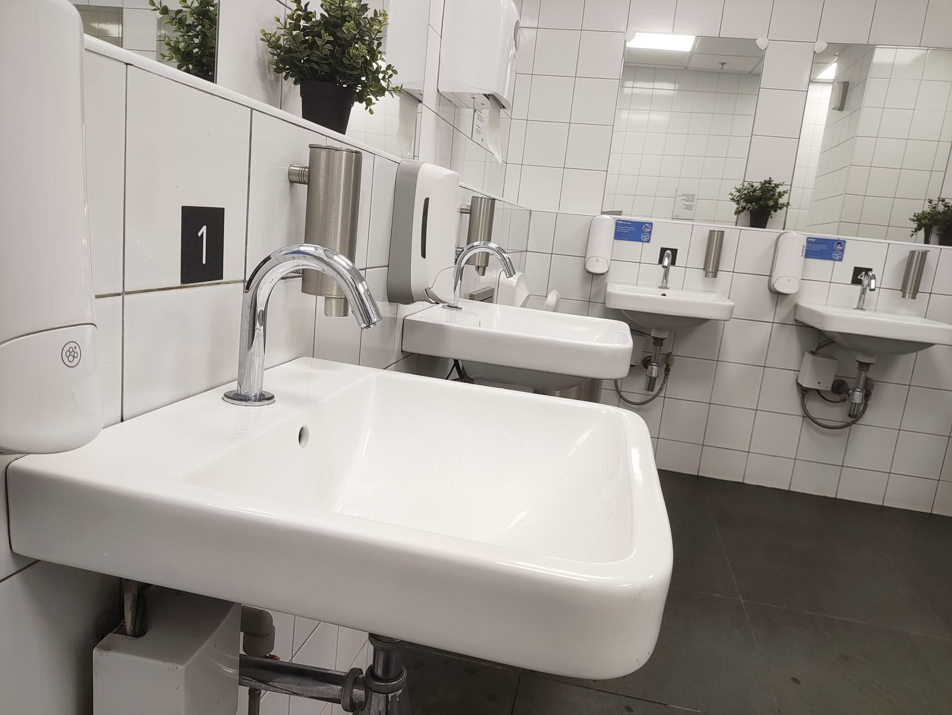 Une rangée de lavabos en céramique blanche avec robinets chromés dans des toilettes publiques carrelées de blanc.