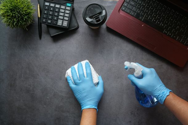 Des mains gantées de gants en nitrile bleus nettoient la surface d'un bureau sombre avec un chiffon et un vaporisateur, près d'un ordinateur portable et d'une calculatrice.