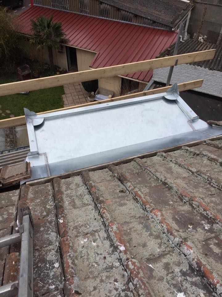 Des solins métalliques sont installés sur un toit de tuiles rouges patinées. Des poutres en bois sont posées au-dessus.