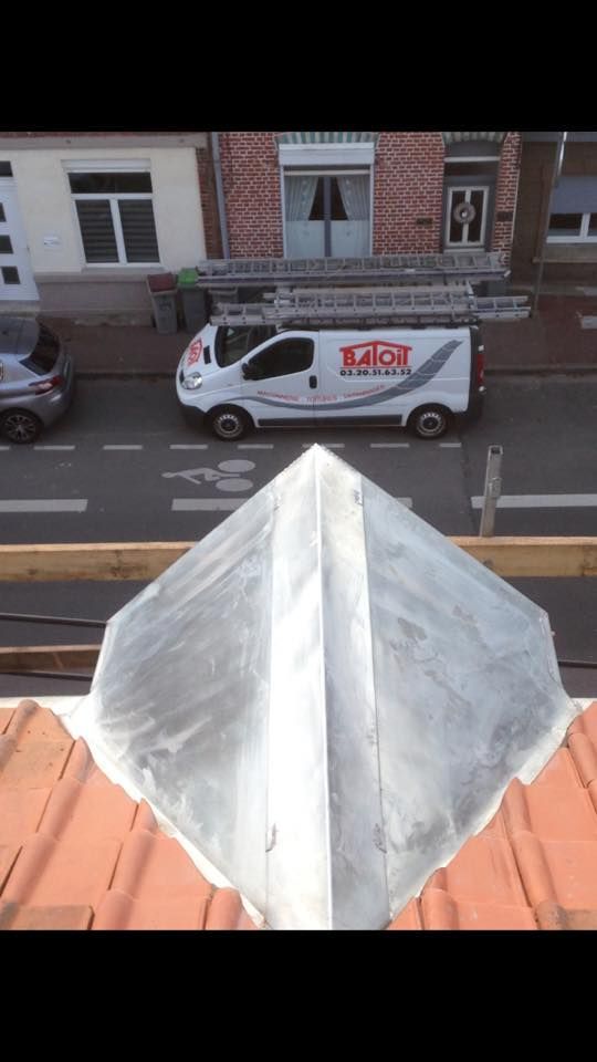 Vue depuis le toit : camionnette blanche avec logo garée dans la rue, ouvrier près d’une échelle, toit de tuiles rouges et bâtiments en briques.