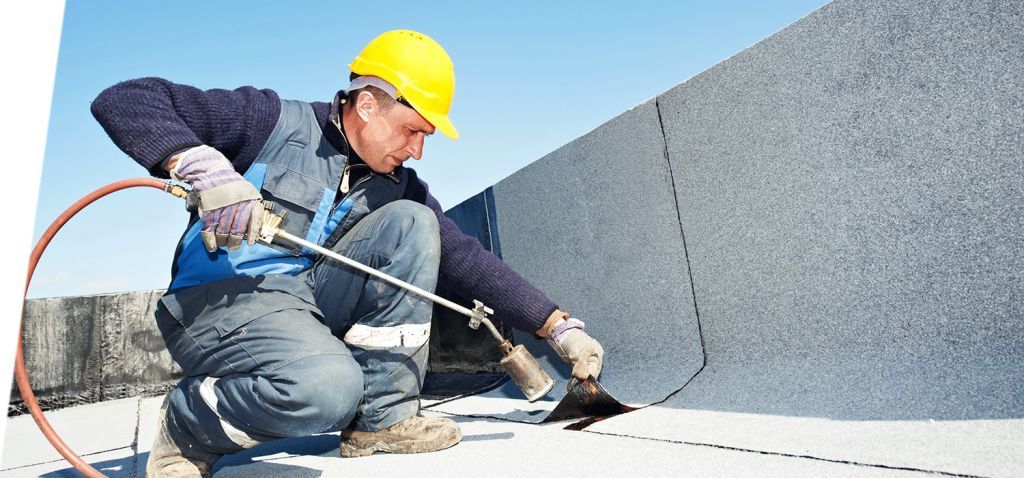 Un ouvrier du bâtiment portant un casque jaune applique du mastic sur un toit plat à l'aide d'un chalumeau.