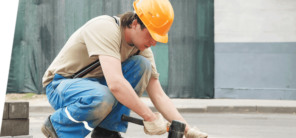 Un ouvrier du bâtiment utilise un marteau, porte un casque de chantier et est agenouillé sur le trottoir.