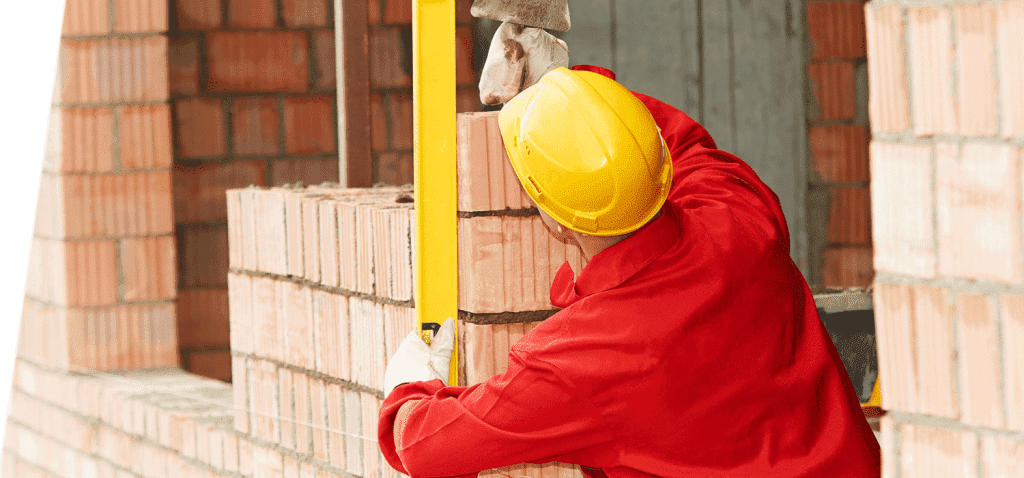Un ouvrier du bâtiment, vêtu d'une combinaison rouge et d'un casque jaune, utilise un niveau sur un mur de briques.