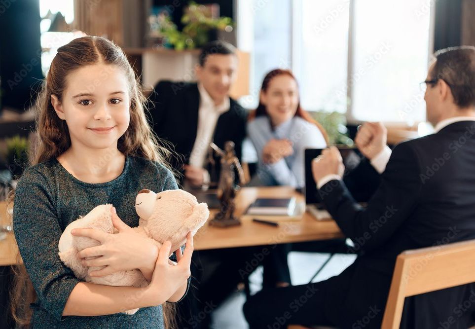 Un enfant tient son nounours et sourit, en arrière plan un homme en costume discute avec son papa et sa maman