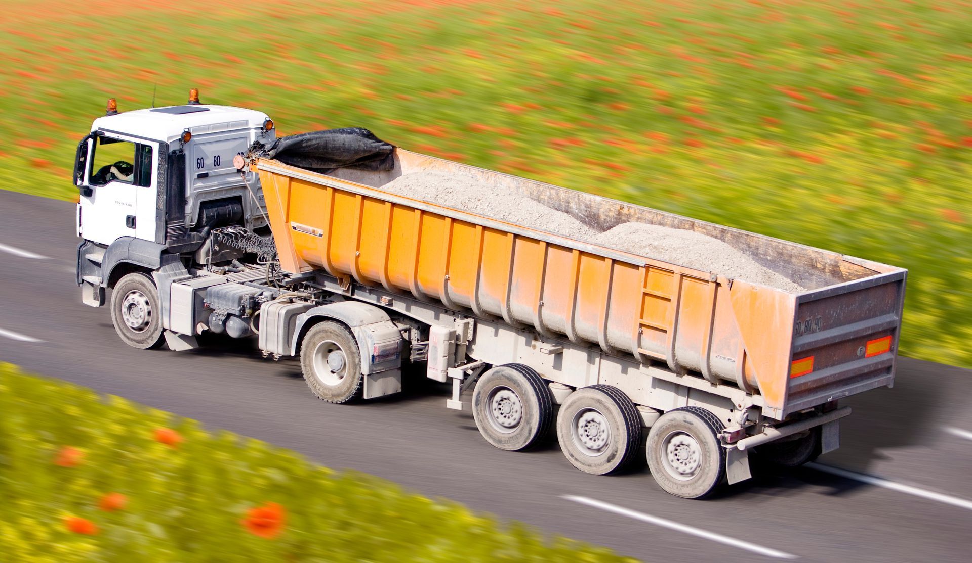 Un camion-benne blanc avec une remorque orange pleine roule sur une route goudronnée.