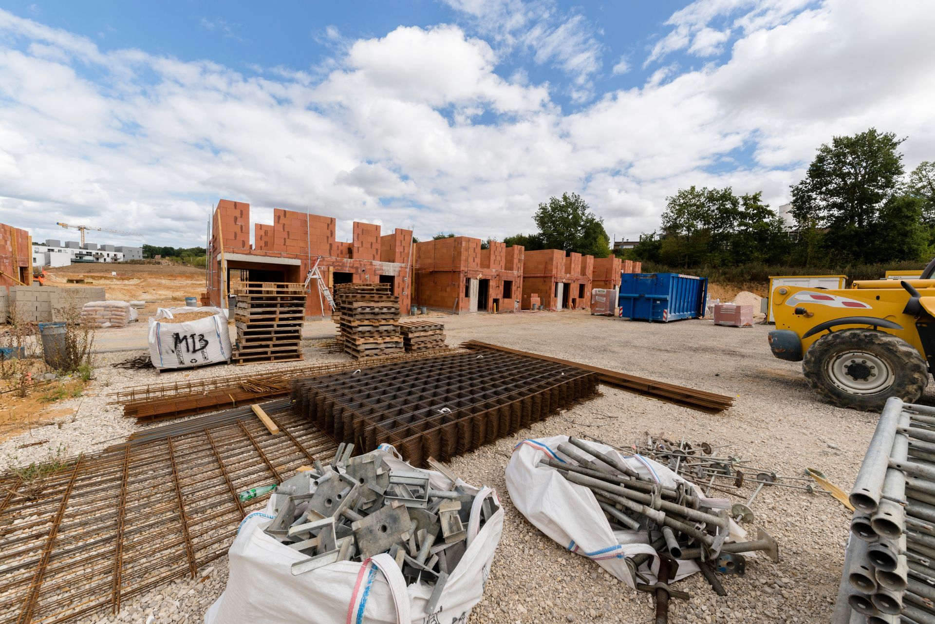 Un chantier de construction avec des structures en briques, des palettes, des piles de barres d'armature, du matériel de construction et des sacs de matériaux de construction.