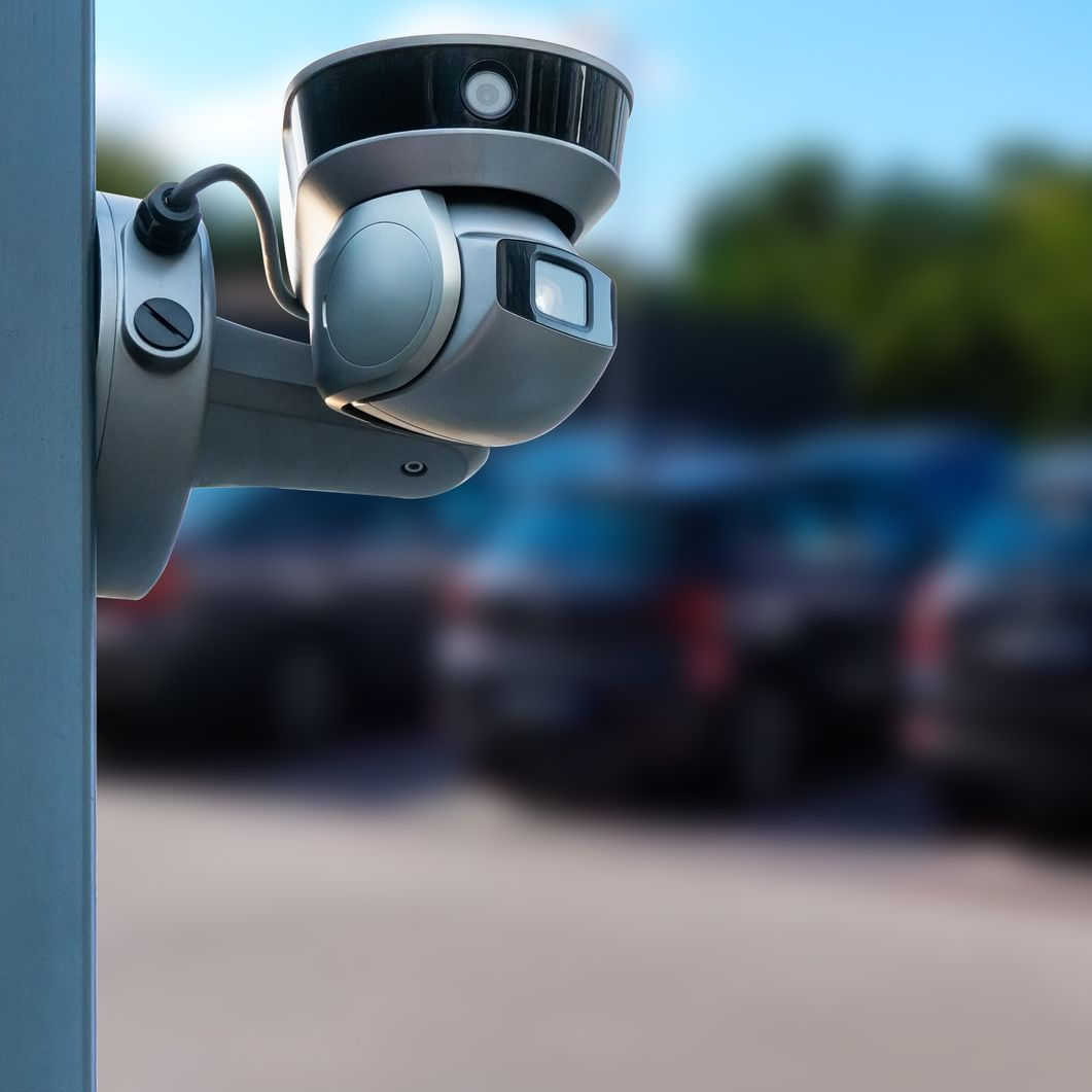 Une caméra de sécurité grise, fixée sur un poteau, surplombe un parking extérieur.