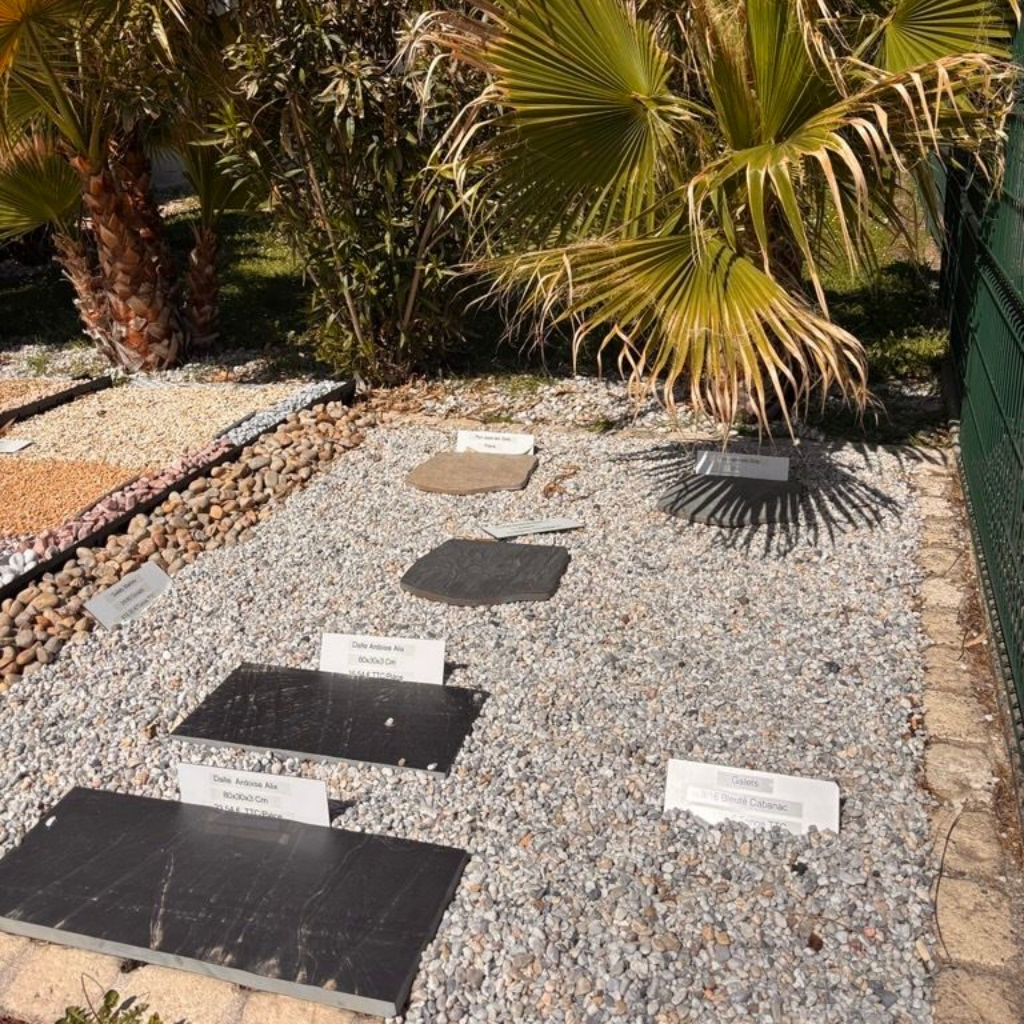 Présentoir extérieur en pierre présentant divers échantillons de carreaux disposés sur un sol de gravier avec des étiquettes.