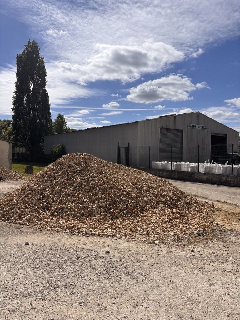 Un grand tas de copeaux de bois se trouve à l'extérieur, à côté d'un entrepôt métallique, sous un ciel bleu partiellement nuageux.