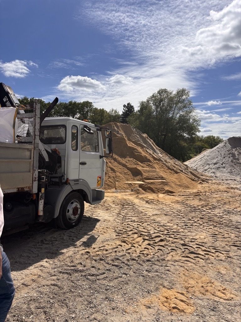 Un camion-benne blanc garé à côté d'un grand tas de sable beige sous un ciel partiellement nuageux.