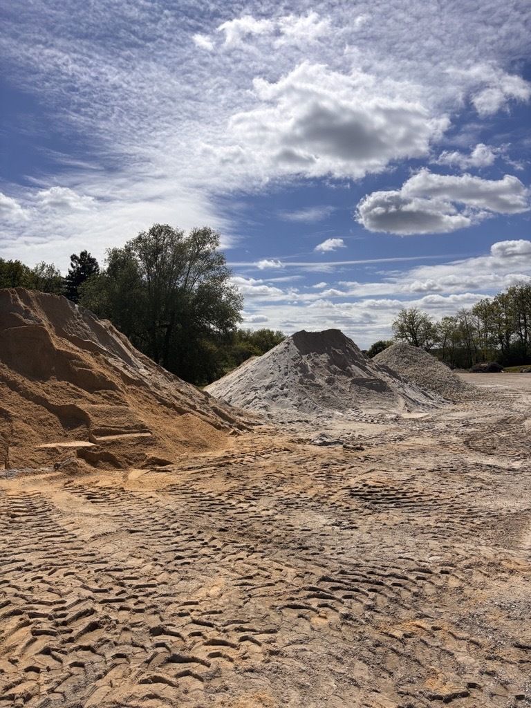 Des tas de sable et de gravier beiges et gris sur un terrain vague sous un ciel bleu parsemé de nuages ​​épars.