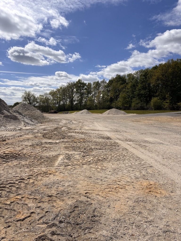Une vaste clairière recouverte de gravier, parsemée de tas de terre, sous un ciel bleu parsemé de nuages ​​blancs, bordée d'une dense rangée d'arbres.