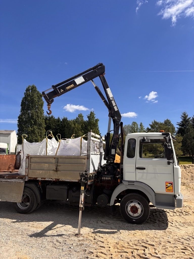Un camion blanc équipé d'une grue et d'un stabilisateur est stationné sur un terrain gravillonné sous un ciel bleu azur.