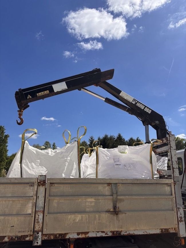 Une grue montée sur le plateau d'un camion, positionnée au-dessus de deux grands sacs blancs remplis.