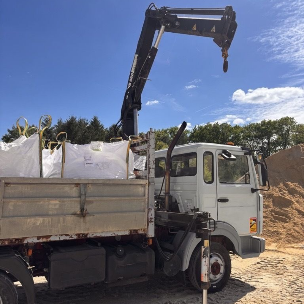 Un camion blanc équipé d'une grue et dont la benne est remplie de grands sacs de rangement blancs est stationné sur un chantier.