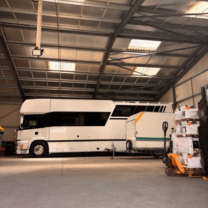 Un camping-car blanc avec une large bande noire est garé à l'intérieur d'un entrepôt, à côté d'une petite remorque et d'un chariot élévateur.