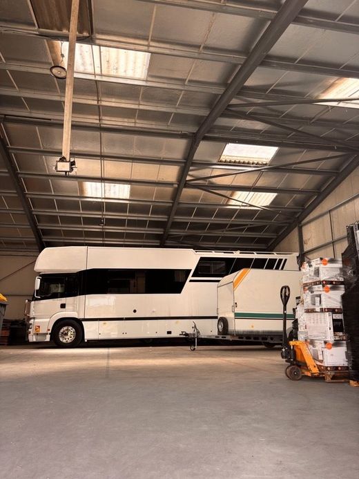 Un grand camping-car blanc est garé à l'intérieur d'un entrepôt spacieux au haut plafond métallique, tirant une remorque plus petite.