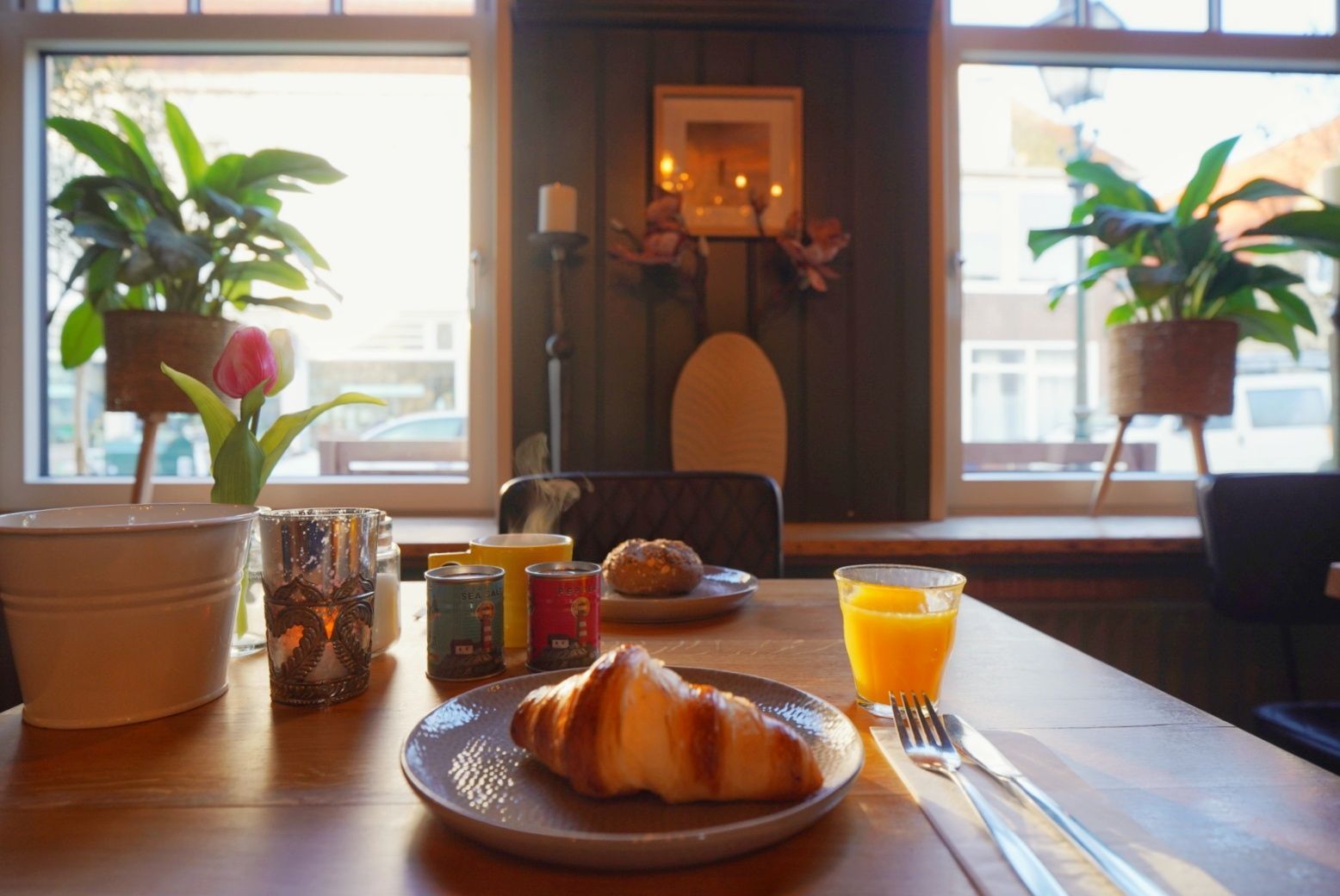 Ontbijttafel met croissant, sinaasappelsap en potplanten bij een licht raam.