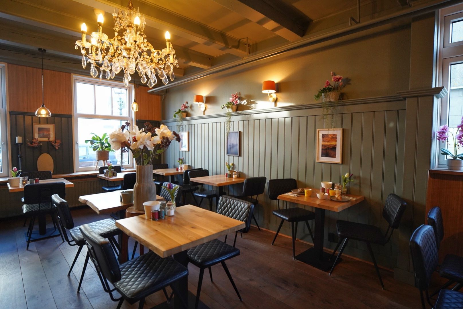 Gezellig café-interieur met houten tafels, kroonluchter, bloemen en zachte sfeerverlichting.
