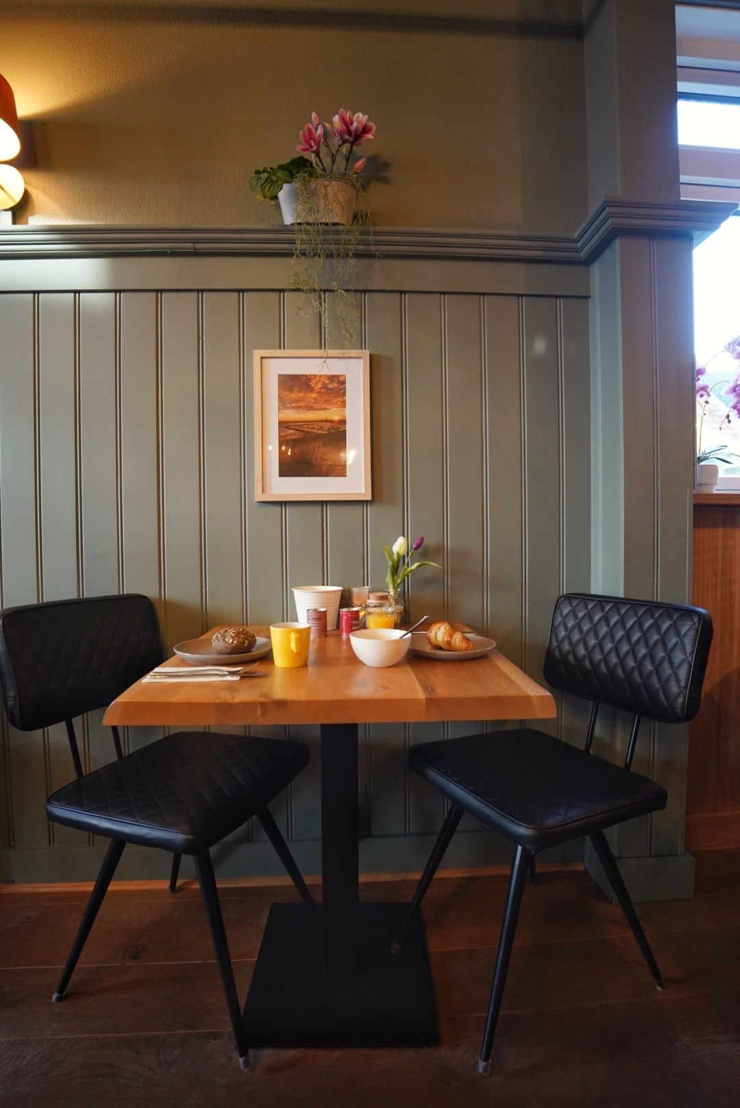 Een klein houten tafeltje tegen een grijze muur, gedekt met mokken en bloemen, tussen twee zwarte stoelen in een gezellig café.
