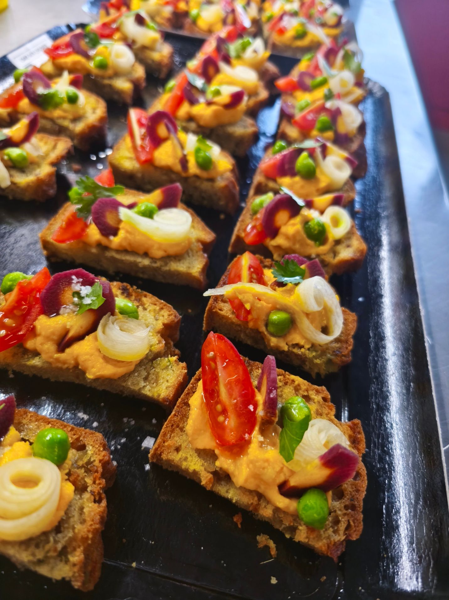 Une personne utilise une spatule jaune pour portionner la pâte sur une surface sombre, près d'une rangée de moules à pâtisserie en métal.