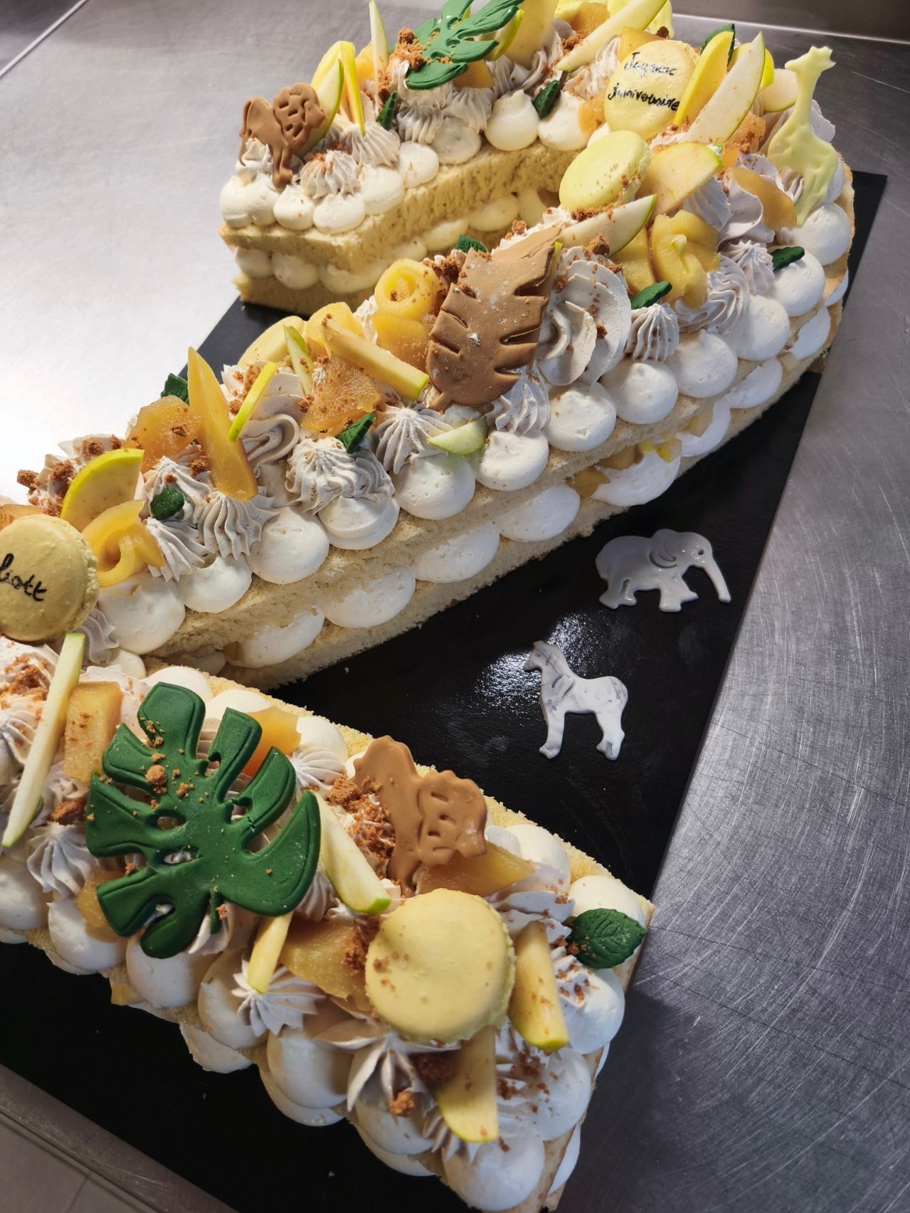 Une pâtisserie rectangulaire à plusieurs couches, décorée de fraises fraîches, de macarons jaunes et de quenelles de meringue vert menthe dressées à la poche à douille.