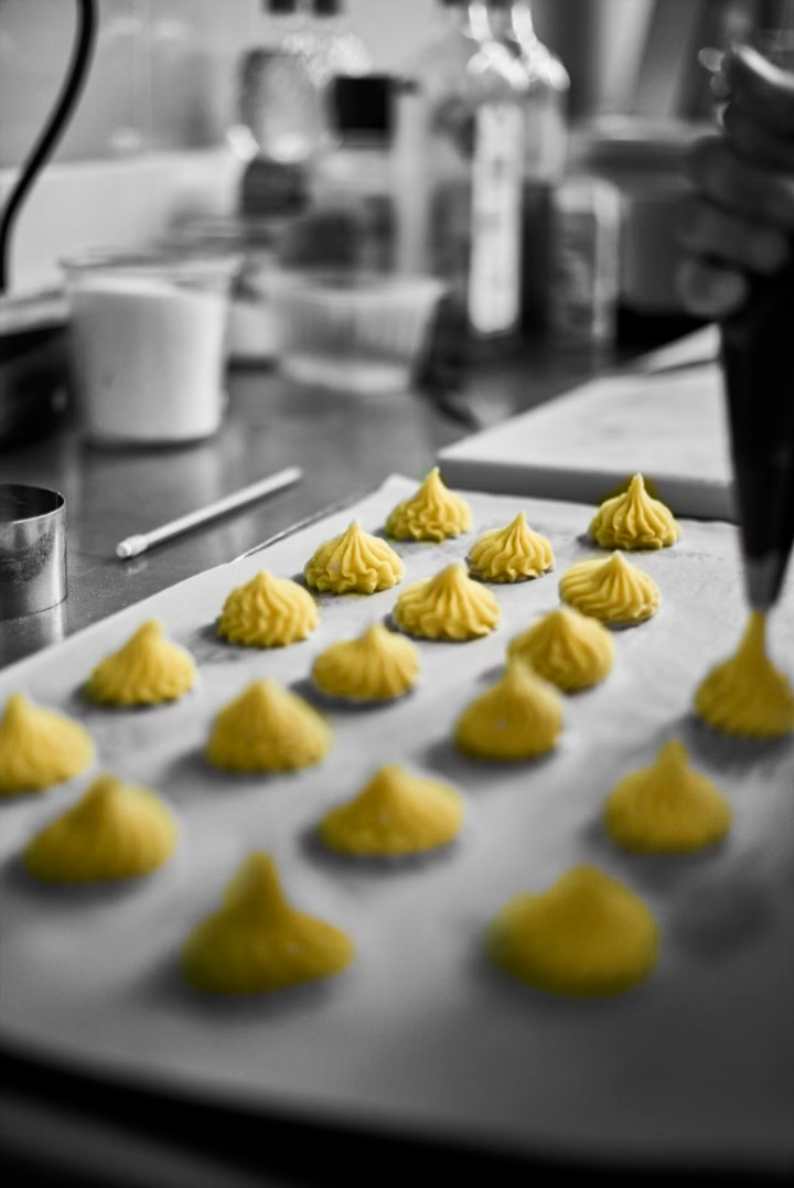 Dans une cuisine, la main d'un chef utilise une poche à douille pour déposer des petites boules de pâte jaune sur une plaque de cuisson recouverte de papier sulfurisé.