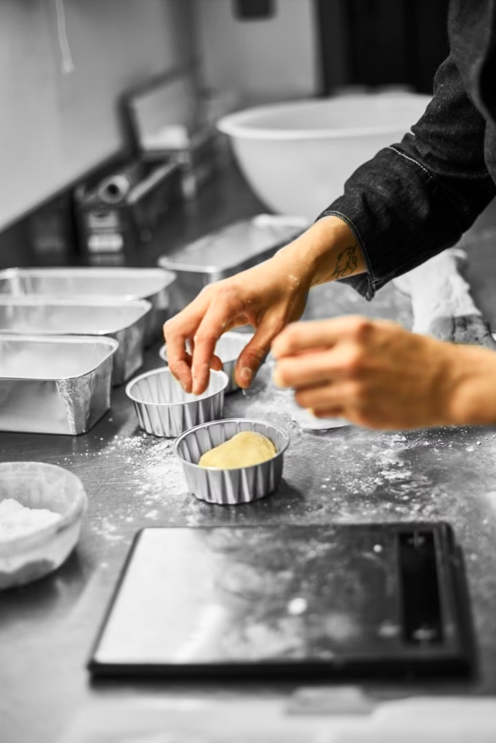 Une personne place un morceau de pâte dans un petit moule à tarte en métal, sur un plan de travail fariné, à côté d'une balance numérique.