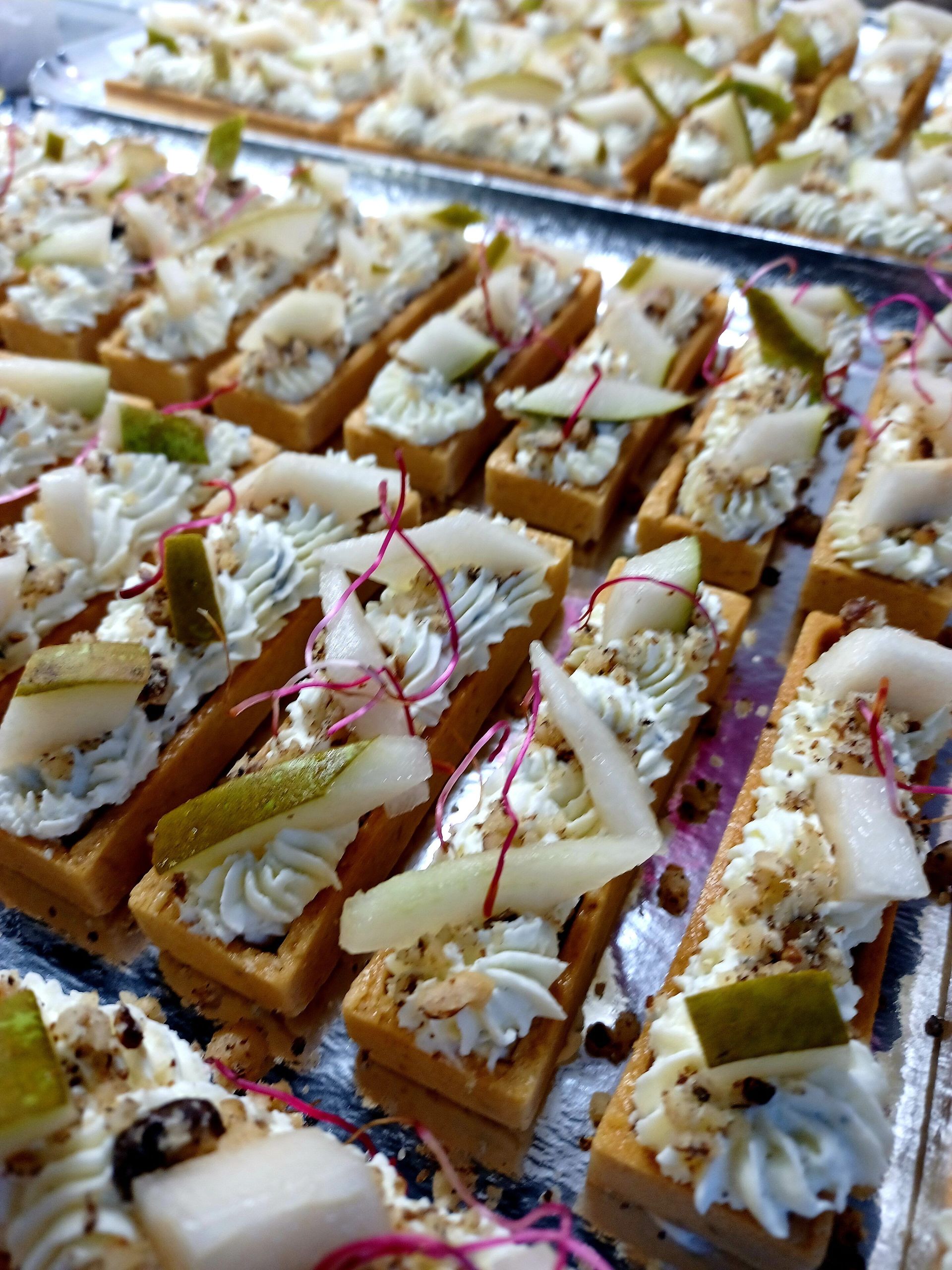 Des rangées de petites tartelettes rectangulaires garnies de crème, de fines tranches de poire et de micro-pousses, disposées sur un plateau de service argenté.