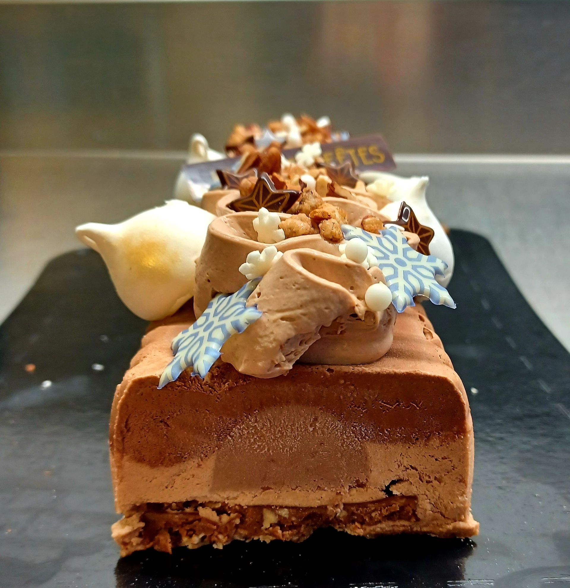 Une bûche de Noël en mousse au chocolat garnie de meringues, de décorations en forme de flocons de neige et de noix, présentée sur un plateau noir.
