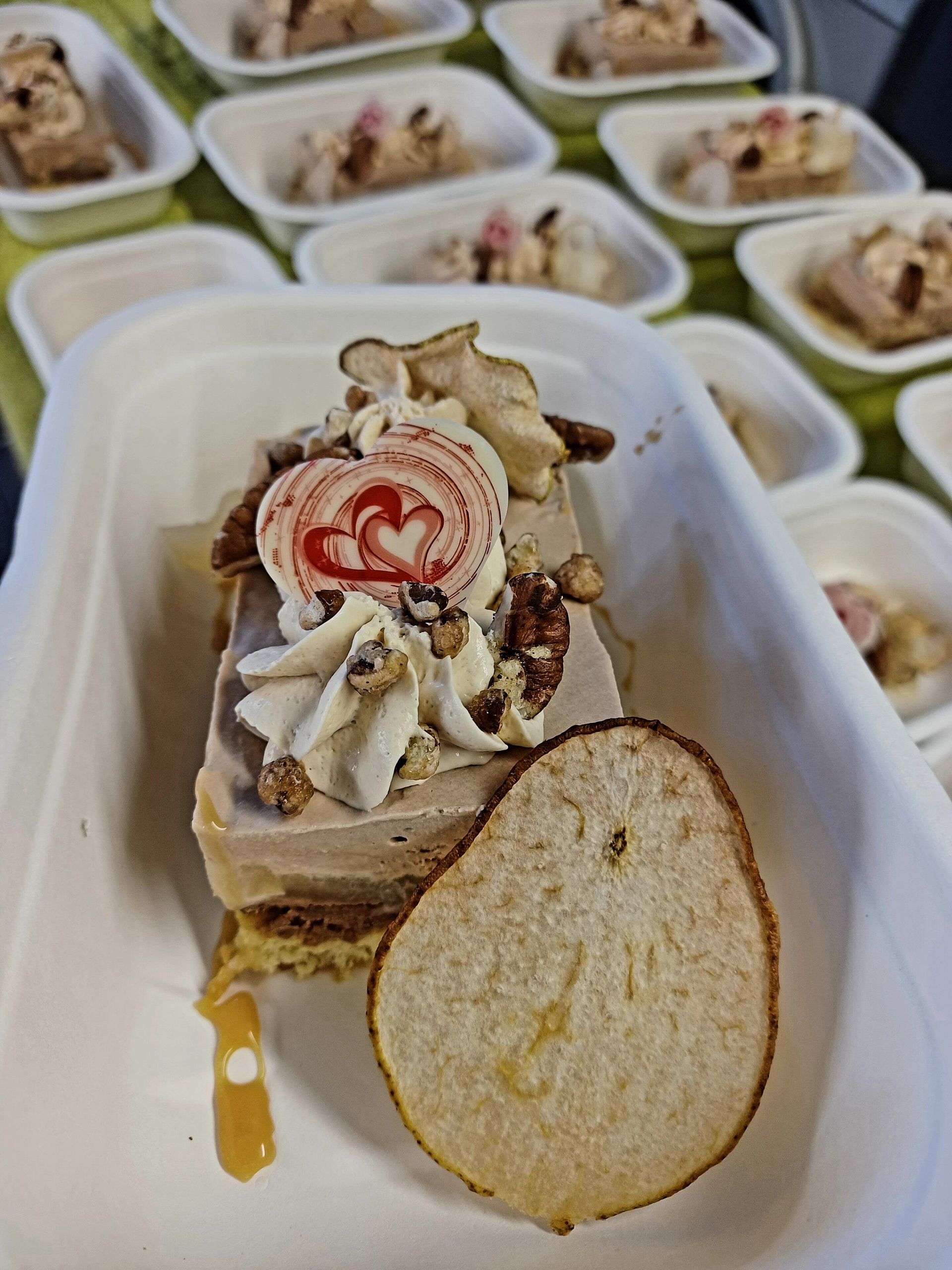 Une portion de gâteau mousse décoré d'un cœur, garnie de crème et d'une tranche de poire séchée, présentée dans un récipient blanc.