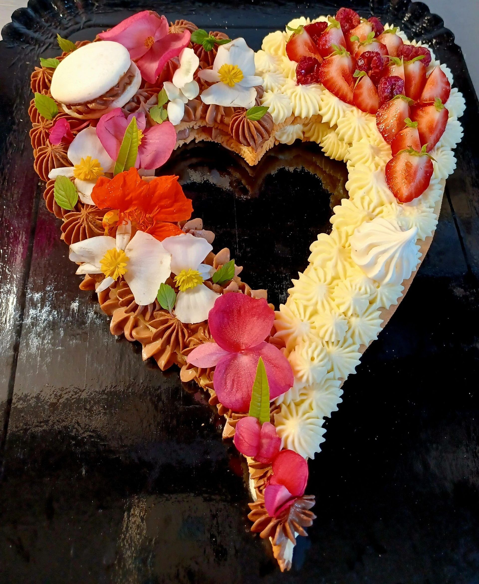 Gâteau en forme de cœur décoré de glaçage, de fleurs, d'un macaron et de fruits rouges frais sur une surface foncée et brillante.