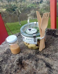 Un bocal en verre contenant de la salade, des couverts en bois et un petit pot de vinaigrette posés sur une souche d'arbre, le tout sur un fond herbeux flou.