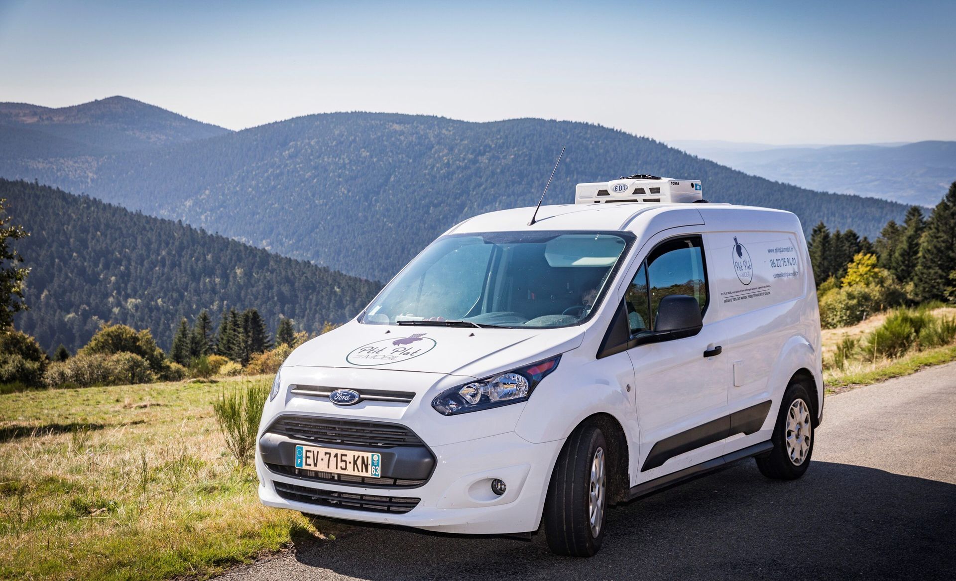 Une camionnette Ford Transit Connect blanche est garée sur une route de montagne, avec des collines ondulantes en arrière-plan.