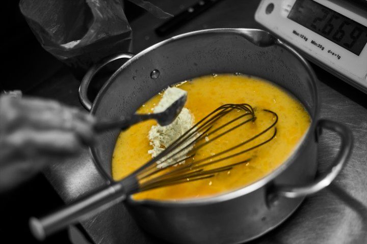 Une main ajoute des ingrédients secs à un liquide jaune dans une casserole en métal, avec un fouet à proximité et une balance numérique visible.