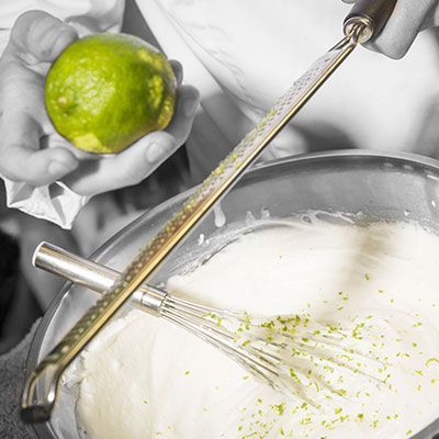 Une personne qui râpe le zeste d'un citron vert dans un bol de pâte crémeuse à l'aide d'un fouet et d'un zesteur en métal.