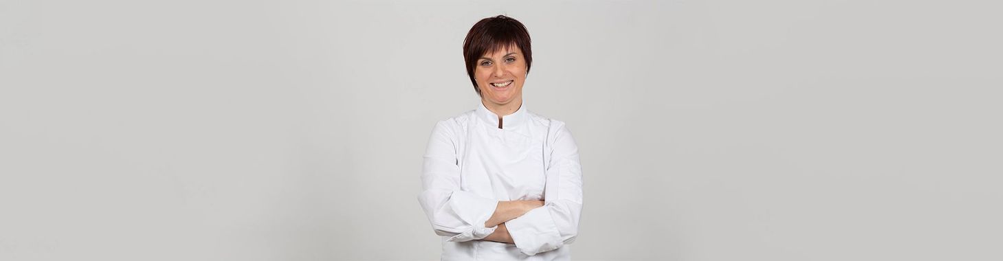 Une personne vêtue d'un uniforme de chef blanc se tient devant un fond uni, les bras croisés, souriante.