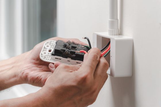 Manos instalando tomacorriente eléctrico; cables negros y rojos en caja blanca en la pared.