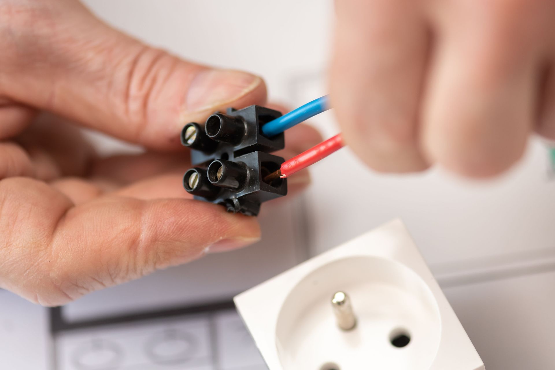Persona que conecta cables rojos y azules a un bloque de terminales negro, con una toma de corriente blanca cerca.
