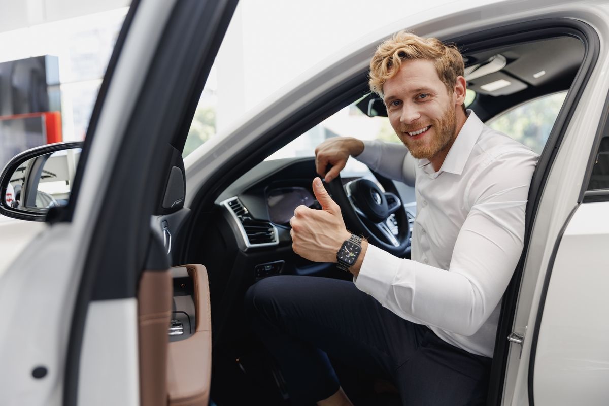Un hombre sonríe sentado en el asiento del conductor de un coche blanco, asomado por la puerta abierta