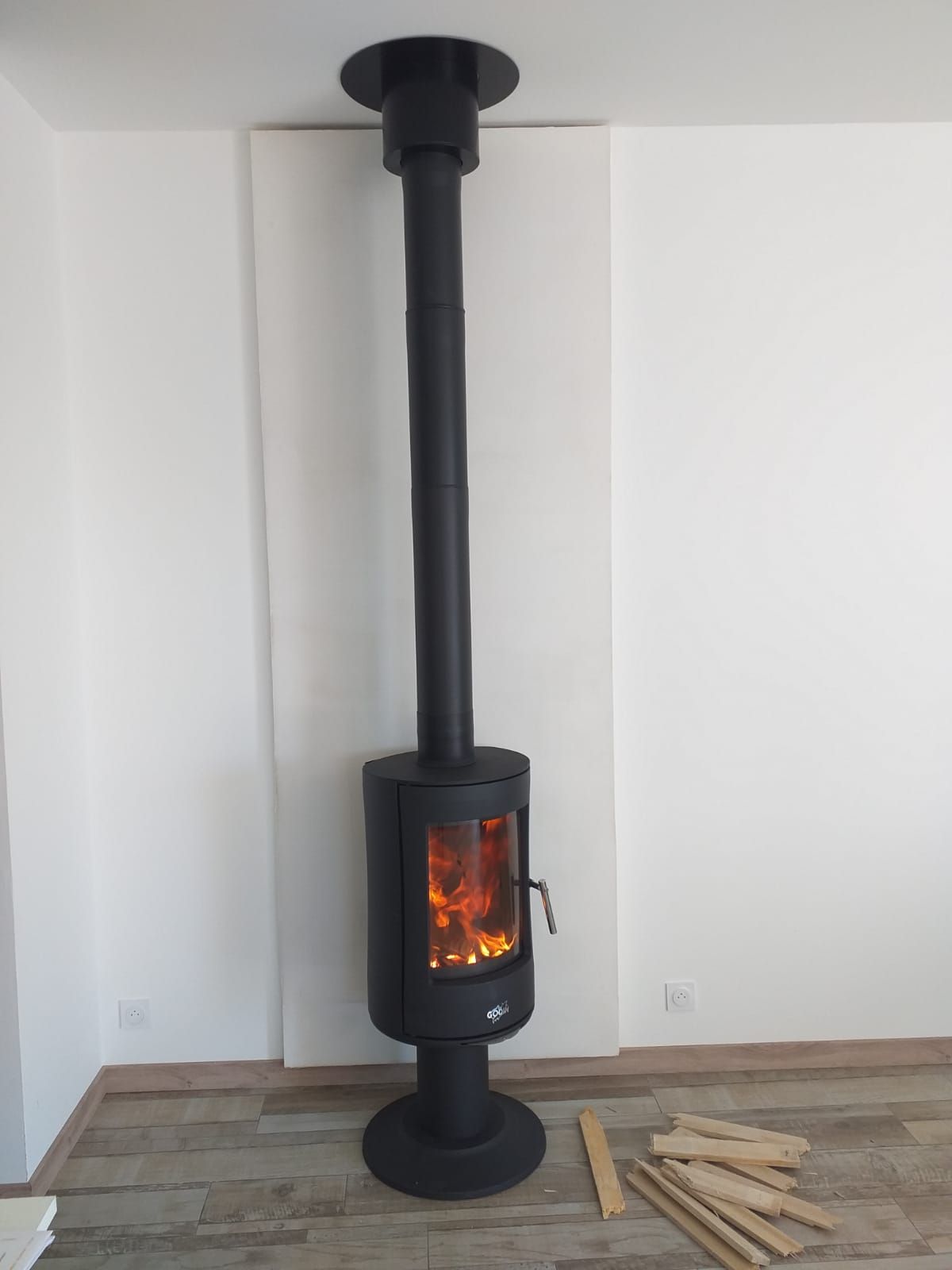 Poêle à bois noir avec feu allumé, relié à une haute cheminée noire, installé dans une pièce aux murs blancs, à côté d'une pile de bois de chauffage.