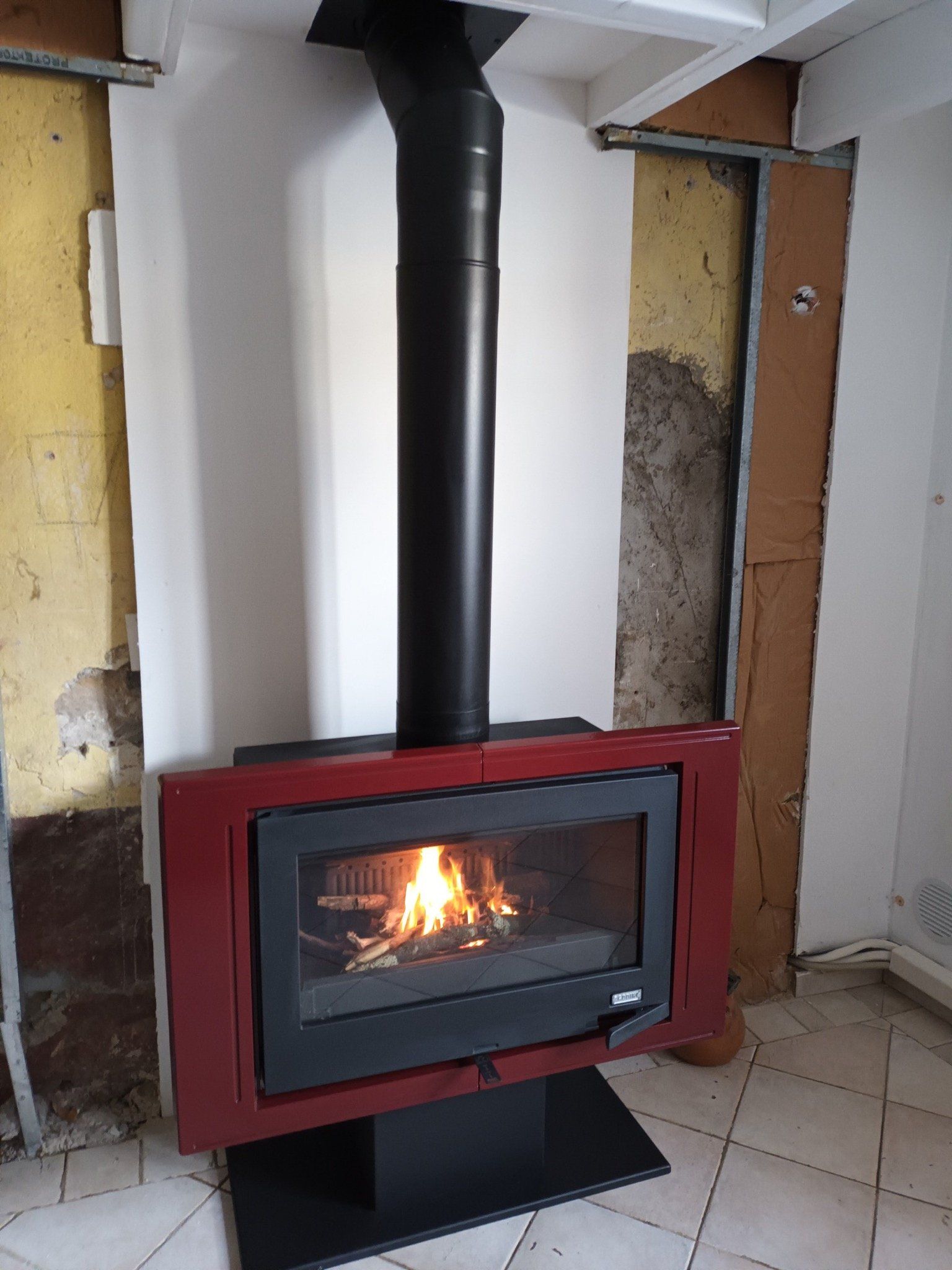 Cheminée rouge avec des flammes qui brûlent, conduit de cheminée noir, mur blanc avec charpente apparente.