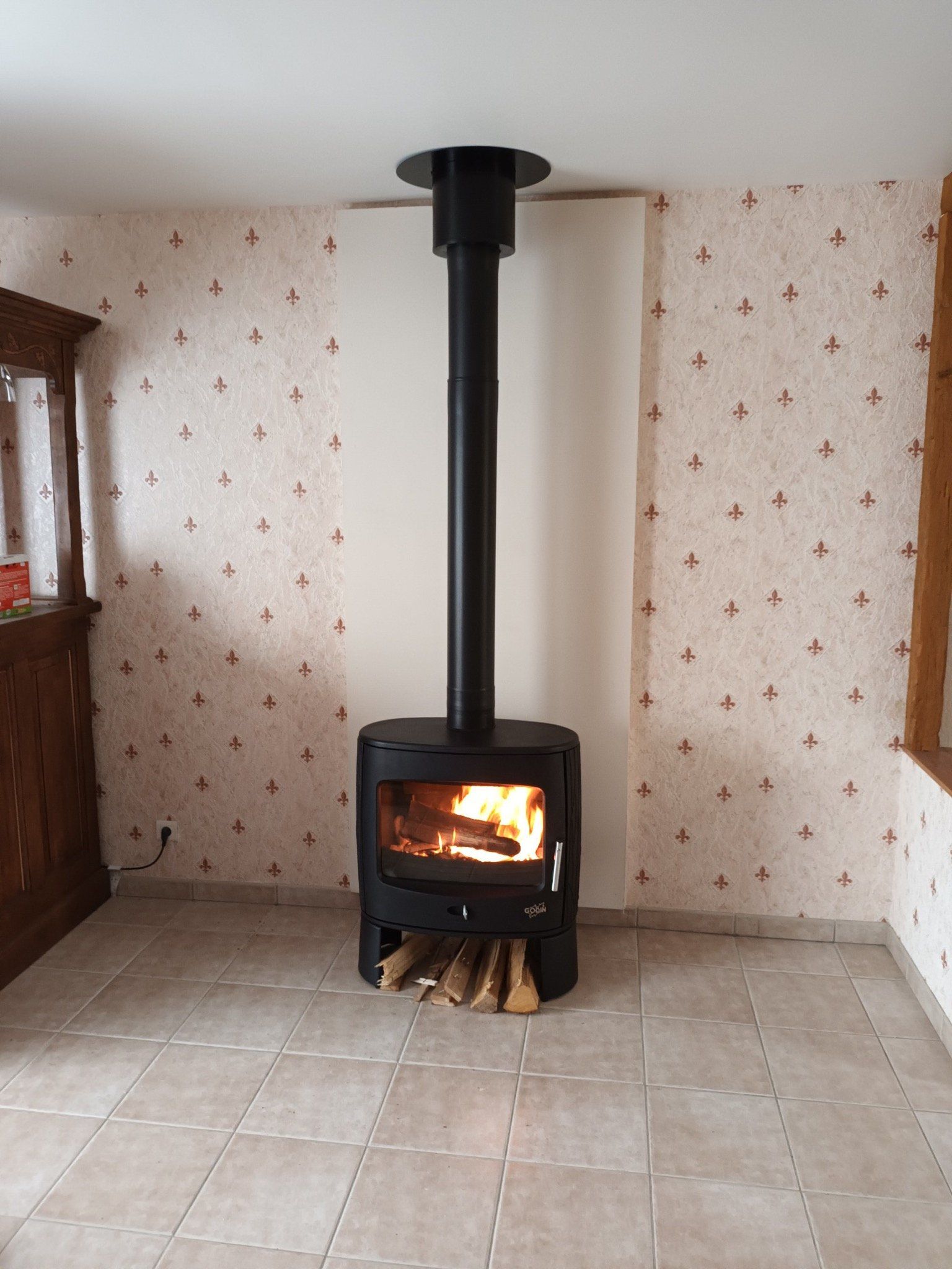 Poêle à bois avec feu allumé et cheminée noire dans une pièce avec papier peint à motifs et sol carrelé beige.