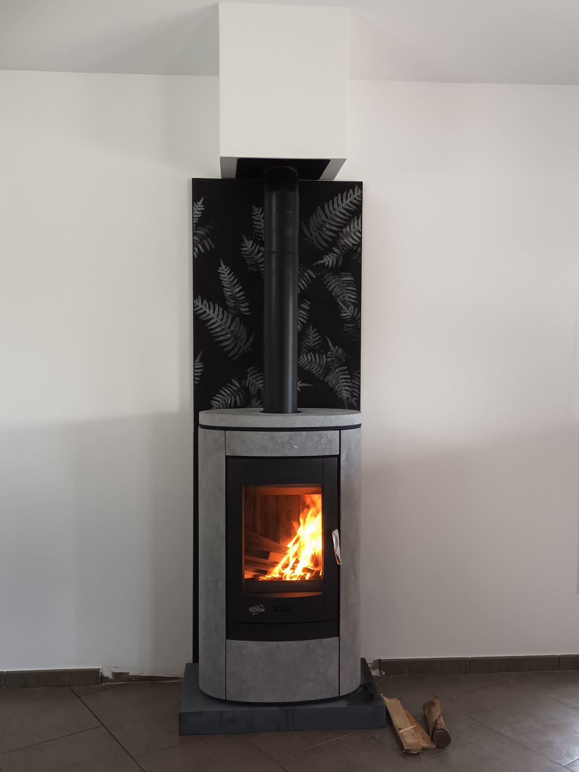Un poêle à bois allumé, avec une crédence à motifs noirs et gris et une cheminée blanche, se détache sur un mur blanc.