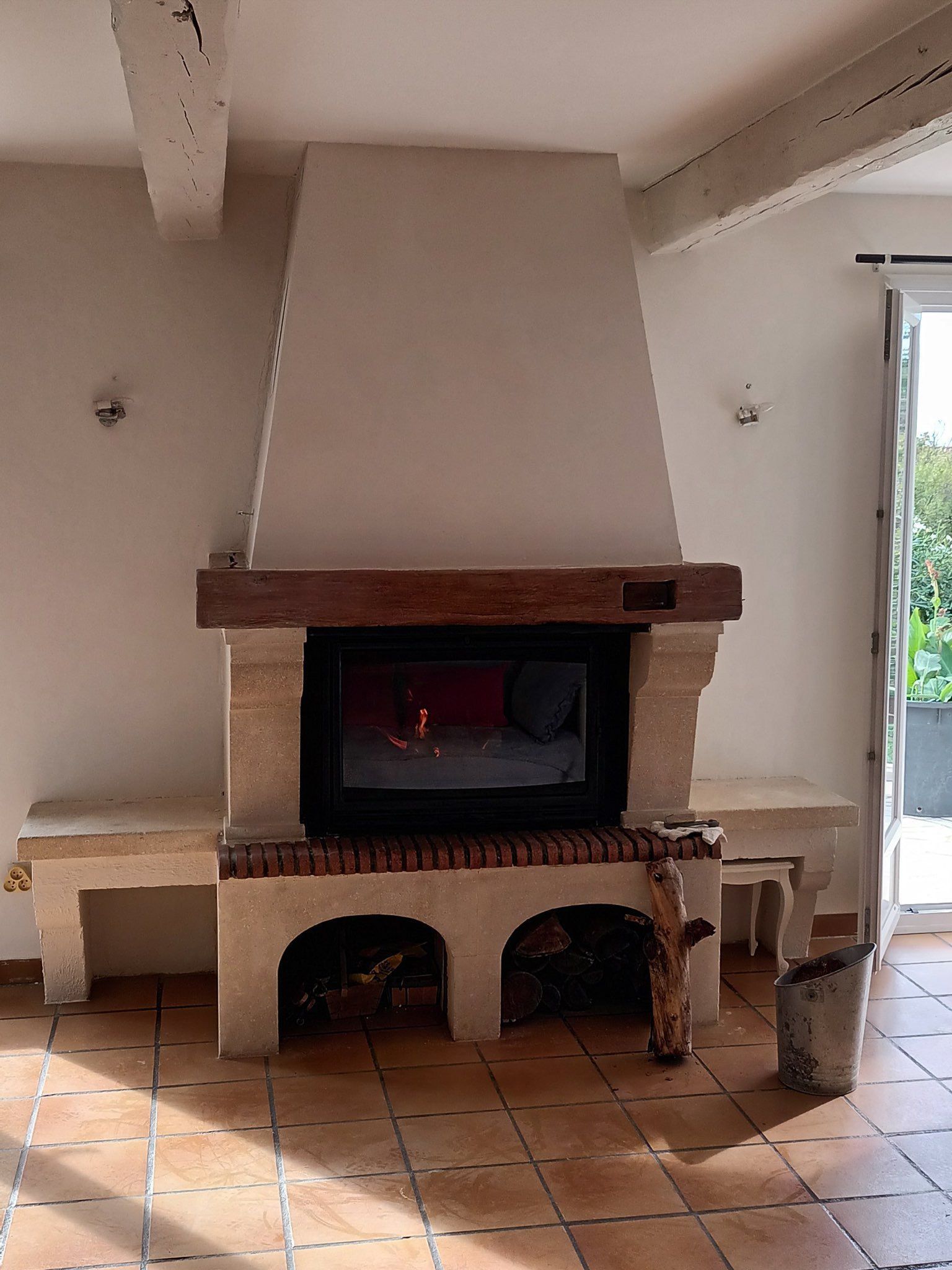 Cheminée avec feu allumé, construction en pierre et en bois, deux bancs intégrés et rangement pour les bûches.