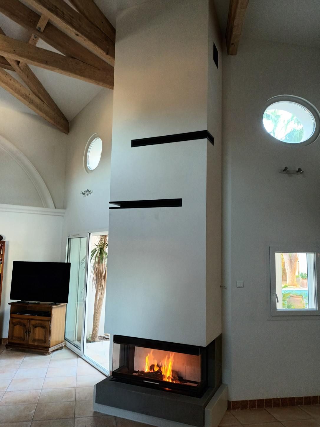 Cheminée moderne à flammes, intégrée à une colonne blanche, dans une pièce lumineuse avec fenêtres.