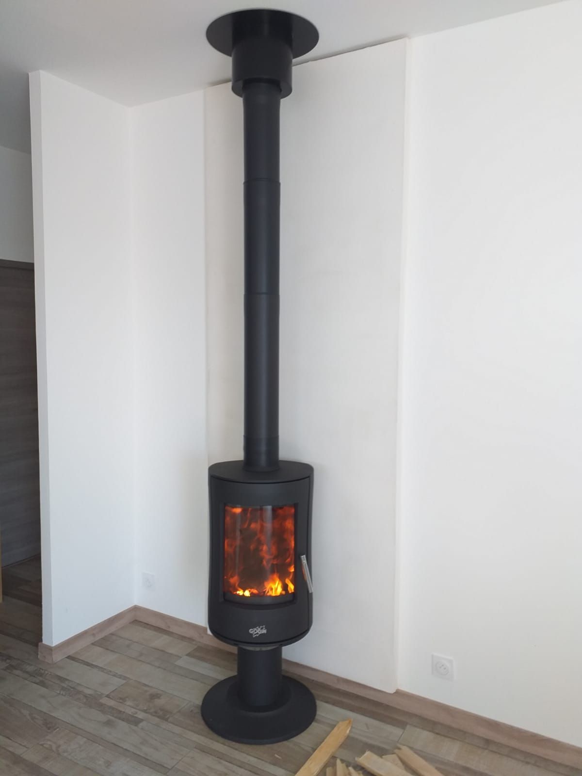 Poêle à bois noir avec feu allumé, relié à une cheminée, placé dans un coin avec des murs blancs et un sol en bois.