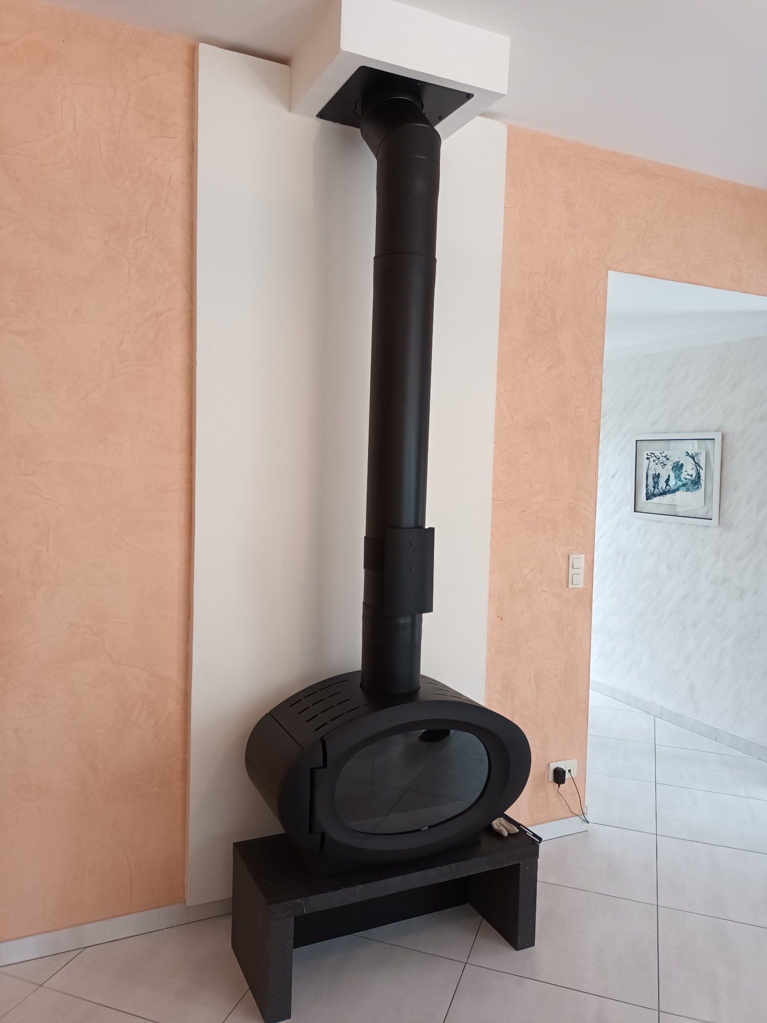 Cheminée noire autoportante à conduit vertical, adossée à un panneau mural blanc sur un mur couleur pêche.