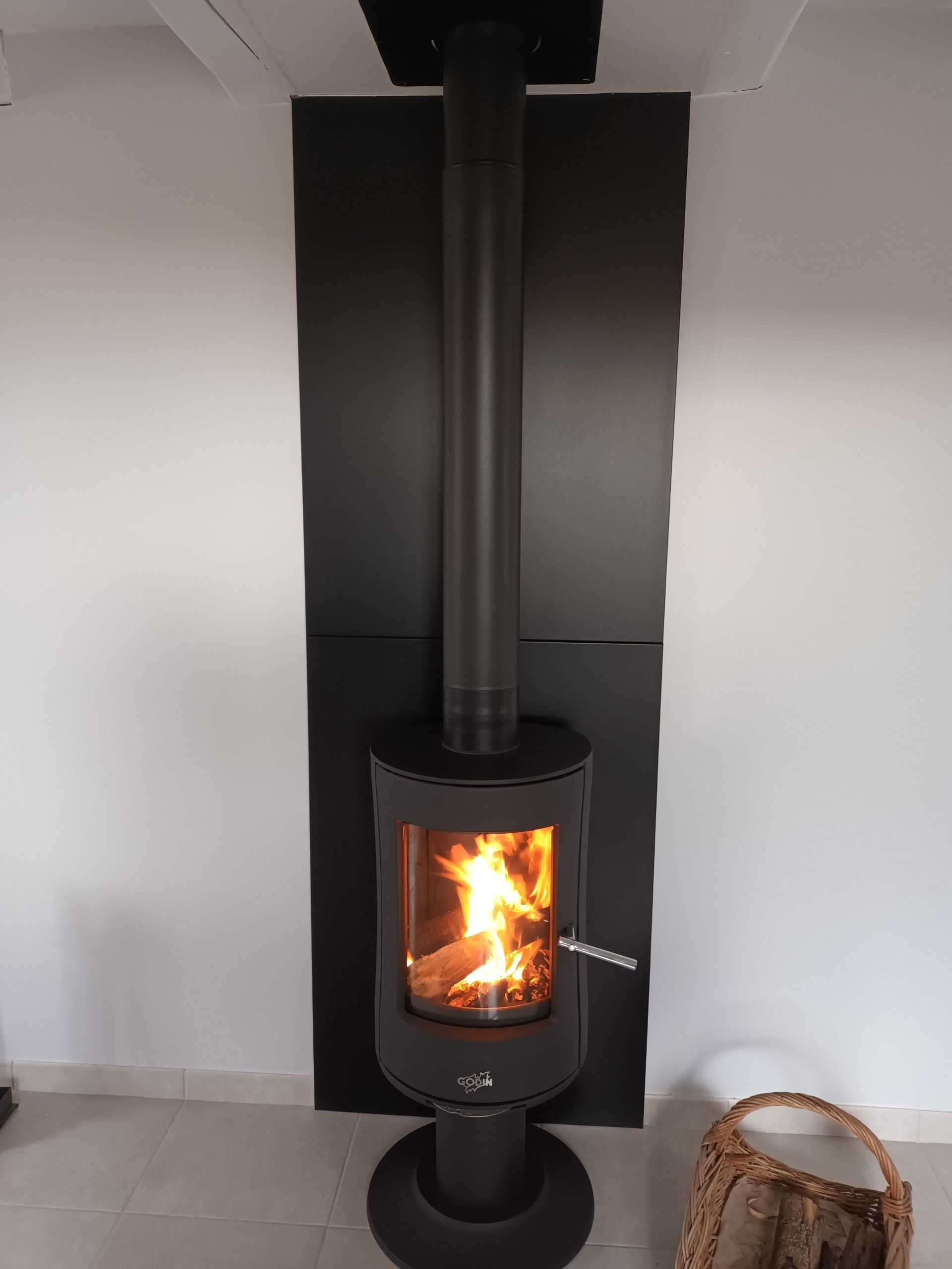 Poêle à bois avec flammes, conduit de cheminée noir, panneau de fond noir sur mur blanc.